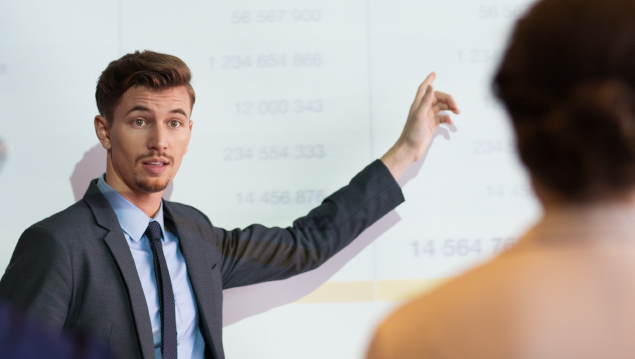 Serious middle-aged businessman standing at projection screen with table and pointing to it while looking and explaining ideas to audience seen partly